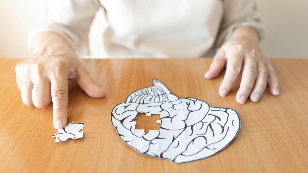 A woman making a jigsaw puzzle about Alzheimer's disease. The jigsaw is of a white brain on a wooden surface. It has one piece missing which is to the side of the brain.