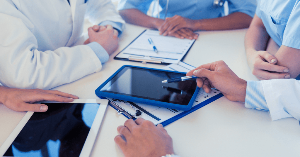 A new care platform: Doctors are sitting around in a circle at a table using tablets and notepads to discuss care management