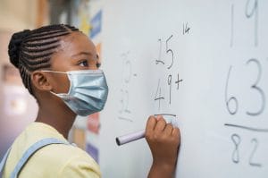 A school child wearing a mask to prevent against long covid writing on a whiteboard with a marker