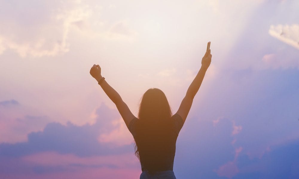 Young empowered lady raising her hands up to the sky, hand holding smart phone against sunset sky ‘Empower women with better Parkinson’s support’