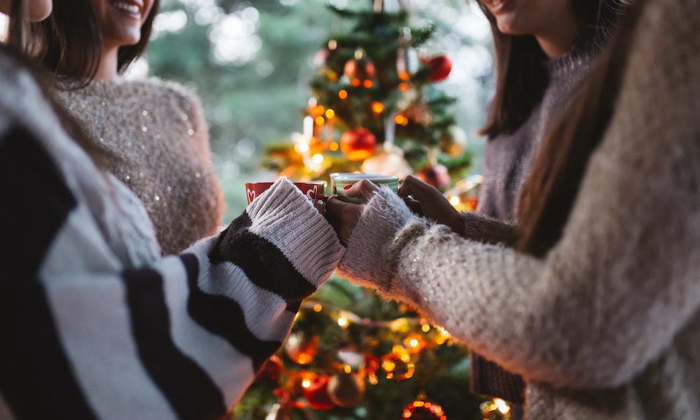 Friend celebrating together and holding cups of warm tea. Christmas tree in background. Four ways Christmas can bring people together